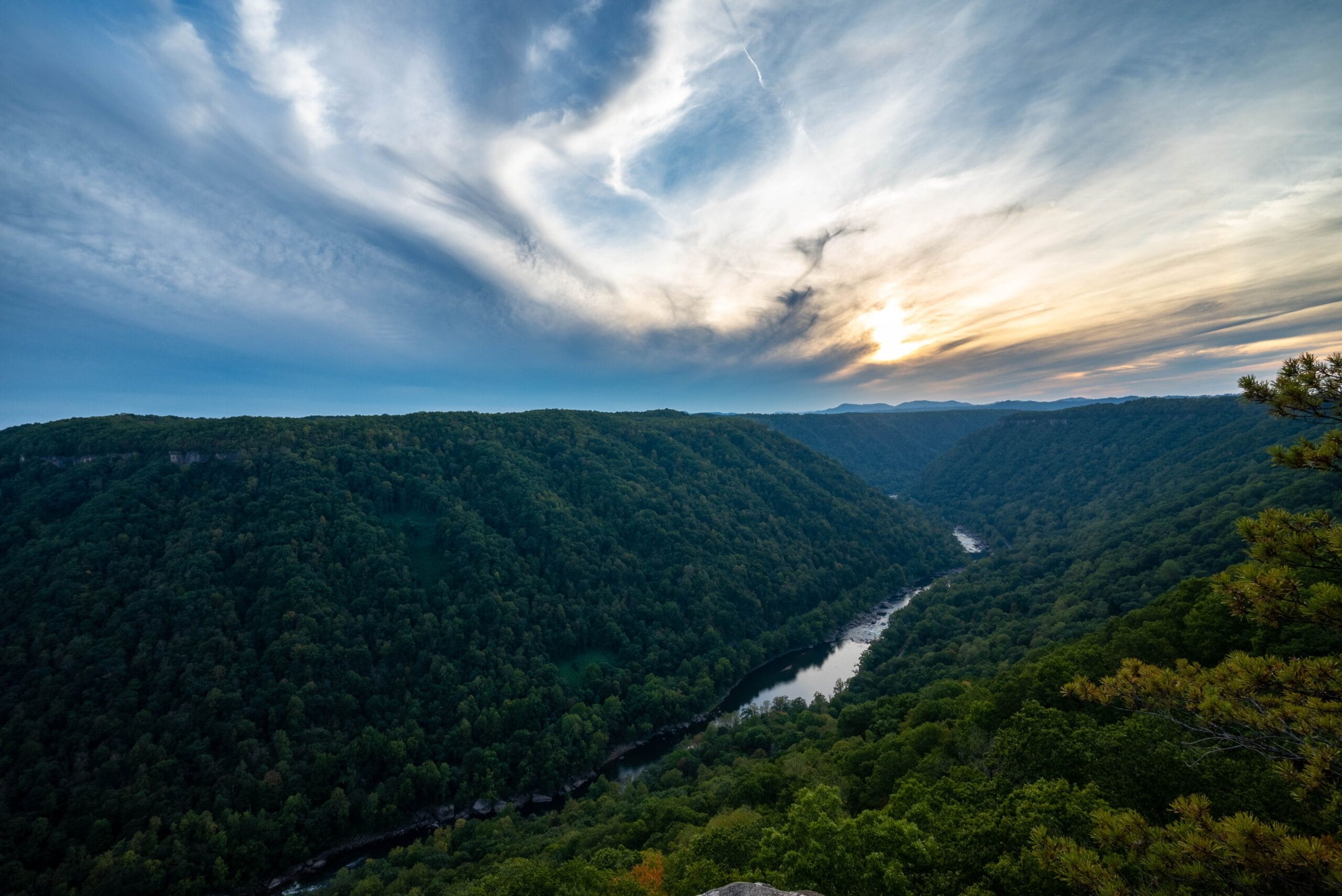 Sunset over a river valley cutting through forested West Virginia ridges, the drainage-and-ridge landscape that shapes how Wetzel County horizontal well units are laid out