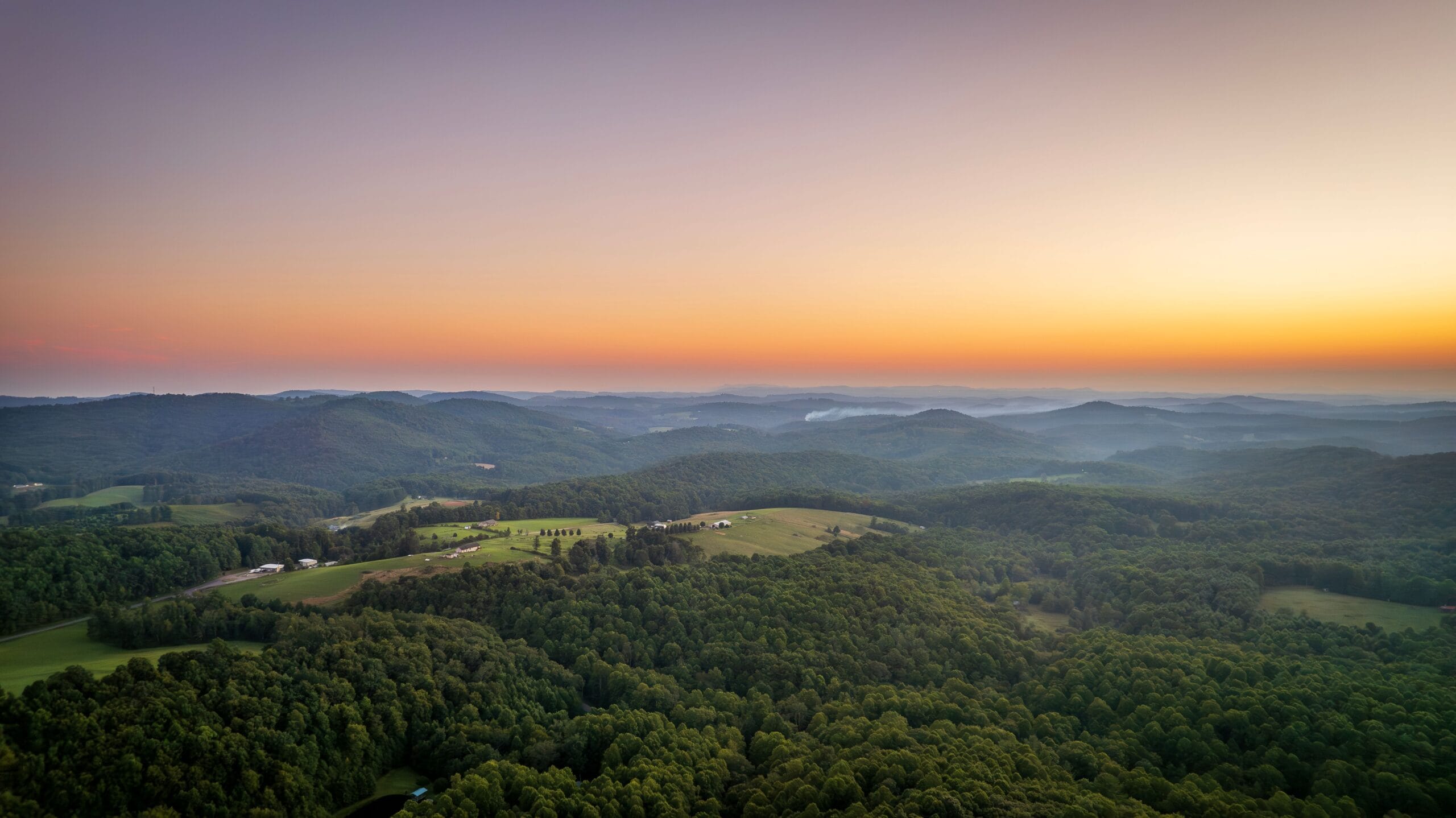 Aerial view at sunrise of forested ridges and scattered hilltop farms, the rolling terrain typical of Doddridge County, West Virginia, where Marcellus laterals run beneath family tracts